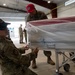 Two military personnel in OCP (Operational Camouflage Pattern) uniforms are working with a flag-draped casket wrapped in protective plastic sheeting inside what appears to be a military hangar. The casket, which displays red, white, and blue colors consistent with an American flag, is positioned on a wheeled transfer case. This appears to be a dignified transfer or mortuary affairs operation.