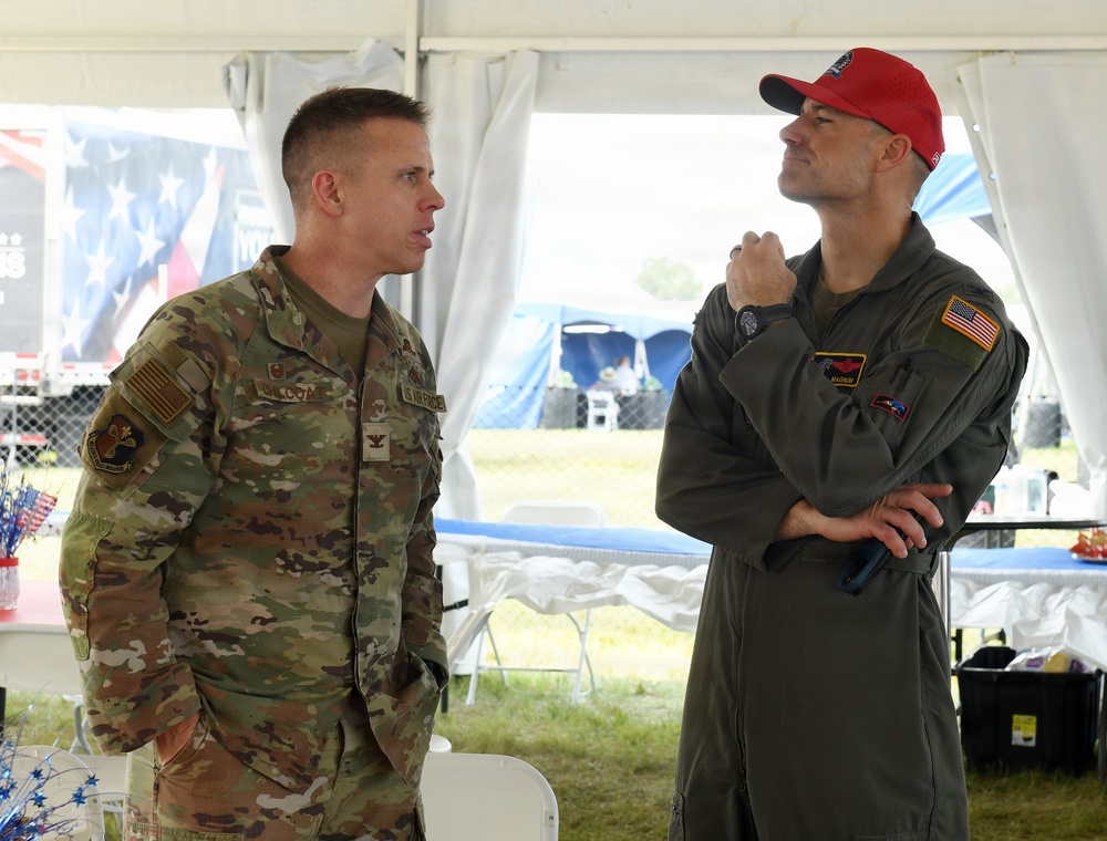 Two U.S. Air Force personnel are conversing under a white tent at what appears to be an outdoor military or patriotic event. The individual on the left wears OCP (Operational Camouflage Pattern) uniform with a Colonel rank insignia, while the individual on the right wears an olive green flight suit with a red cap and holds a microphone. An American flag banner and patriotic decorations are visible in the background.