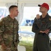 Two U.S. Air Force personnel are conversing under a white tent at what appears to be an outdoor military or patriotic event. The individual on the left wears OCP (Operational Camouflage Pattern) uniform with a Colonel rank insignia, while the individual on the right wears an olive green flight suit with a red cap and holds a microphone. An American flag banner and patriotic decorations are visible in the background.