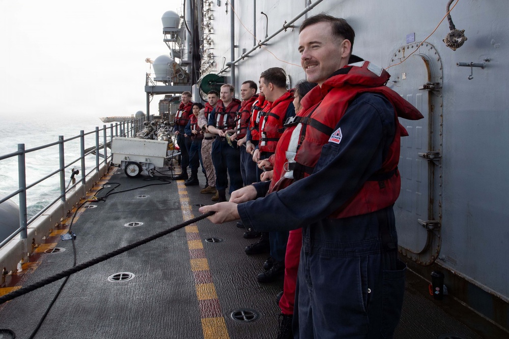 A group of military personnel standing on the deck of a naval vessel at sea, most wearing red life vests over navy blue coveralls. They appear to be engaged in a line-handling or replenishment-at-sea (RAS) evolution, with the foreground figure holding a rope/line. The overcast conditions and open ocean are visible in the background.