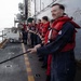 A group of military personnel standing on the deck of a naval vessel at sea, most wearing red life vests over navy blue coveralls. They appear to be engaged in a line-handling or replenishment-at-sea (RAS) evolution, with the foreground figure holding a rope/line. The overcast conditions and open ocean are visible in the background.