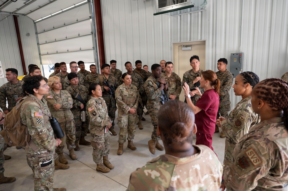 A large group of U.S. Army soldiers in OCP (Operational Camouflage Pattern) uniforms are gathered inside a metal hangar or warehouse facility. Two individuals in maroon/burgundy scrubs appear to be briefing or instructing the assembled soldiers. The setting suggests a training event, medical briefing, or facility tour.