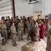 A large group of U.S. Army soldiers in OCP (Operational Camouflage Pattern) uniforms are gathered inside a metal hangar or warehouse facility. Two individuals in maroon/burgundy scrubs appear to be briefing or instructing the assembled soldiers. The setting suggests a training event, medical briefing, or facility tour.