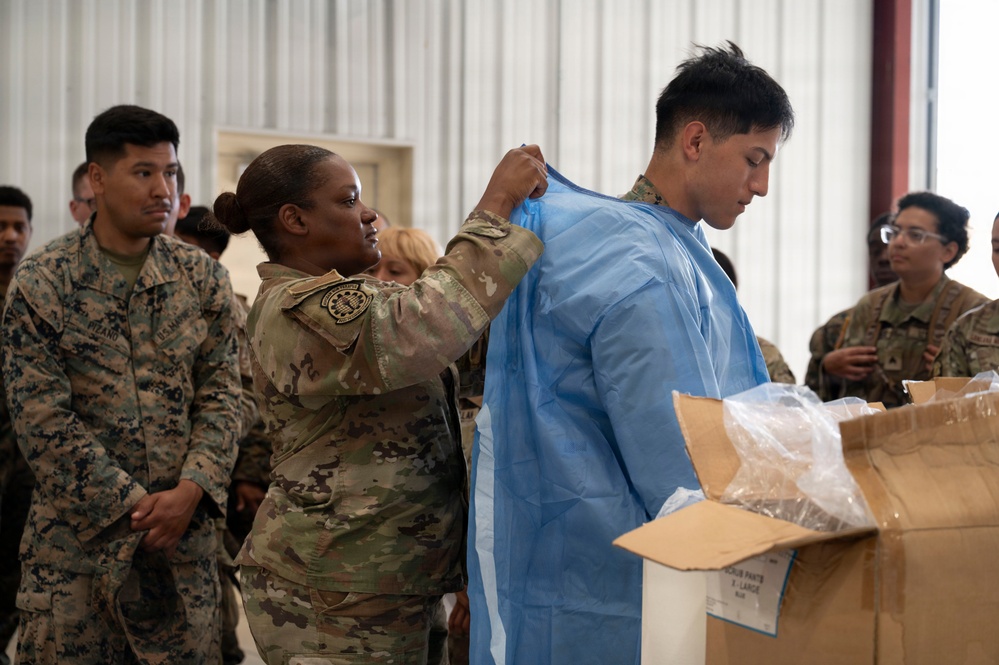 A military training or demonstration scene inside what appears to be a hangar or large facility. A soldier in Army OCP (Operational Camouflage Pattern) uniform is helping another service member don a blue medical/protective gown, while a group of mixed military personnel — including U.S. Marines and Army soldiers — observe. The scene suggests a medical or CBRN (Chemical, Biological, Radiological, Nuclear) protective equipment training exercise.