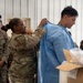 A military training or demonstration scene inside what appears to be a hangar or large facility. A soldier in Army OCP (Operational Camouflage Pattern) uniform is helping another service member don a blue medical/protective gown, while a group of mixed military personnel — including U.S. Marines and Army soldiers — observe. The scene suggests a medical or CBRN (Chemical, Biological, Radiological, Nuclear) protective equipment training exercise.