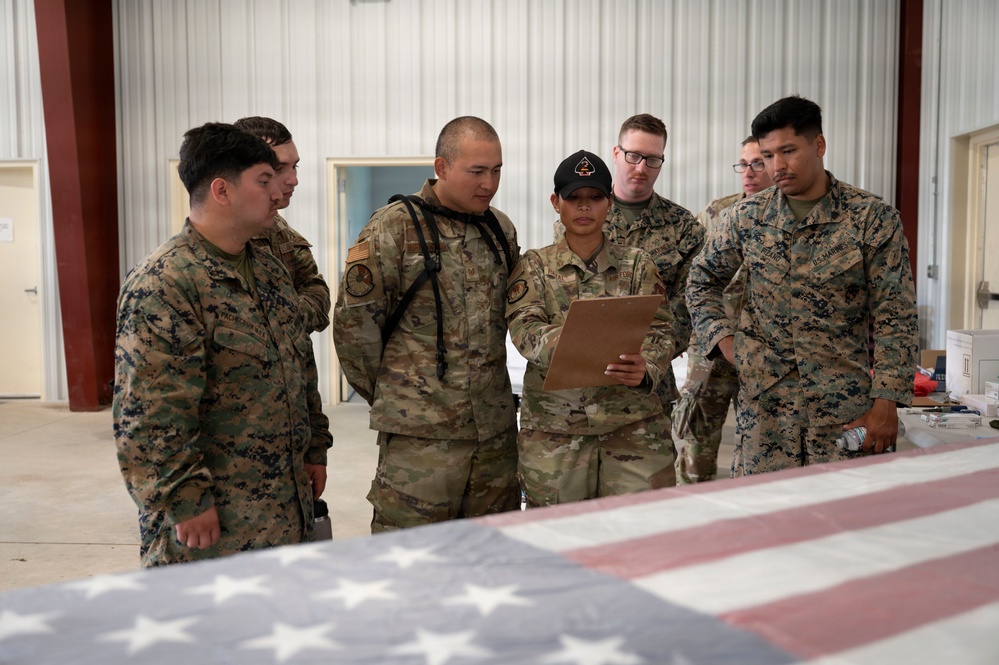 A group of six U.S. military personnel from mixed branches are gathered in what appears to be a hangar or warehouse facility. A central figure in OCP (Operational Camouflage Pattern) uniform is holding a clipboard that others are reviewing. An American flag is draped over a surface in the foreground, suggesting a formal or ceremonial context.