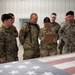 A group of six U.S. military personnel from mixed branches are gathered in what appears to be a hangar or warehouse facility. A central figure in OCP (Operational Camouflage Pattern) uniform is holding a clipboard that others are reviewing. An American flag is draped over a surface in the foreground, suggesting a formal or ceremonial context.