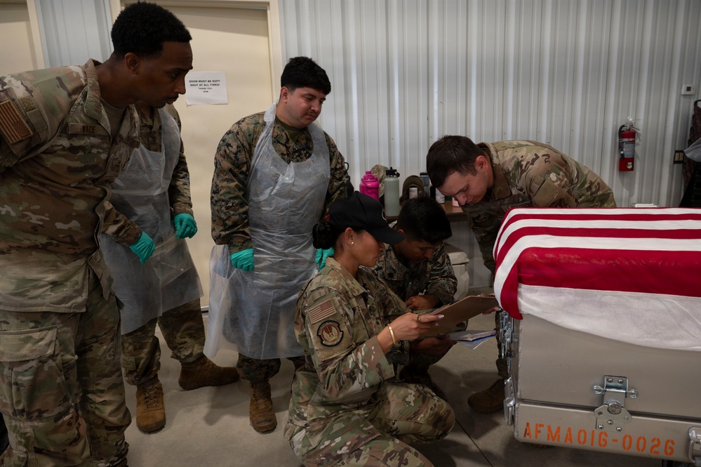 Military personnel from multiple branches are gathered around a flag-draped transfer case or casket marked 'AFMA016-0026', suggesting a mortuary affairs training exercise or dignified transfer procedure. Several service members are wearing protective gear including gloves and plastic aprons, while one is kneeling and writing on a clipboard. The setting appears to be an indoor military facility.