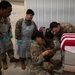 Military personnel from multiple branches are gathered around a flag-draped transfer case or casket marked 'AFMA016-0026', suggesting a mortuary affairs training exercise or dignified transfer procedure. Several service members are wearing protective gear including gloves and plastic aprons, while one is kneeling and writing on a clipboard. The setting appears to be an indoor military facility.