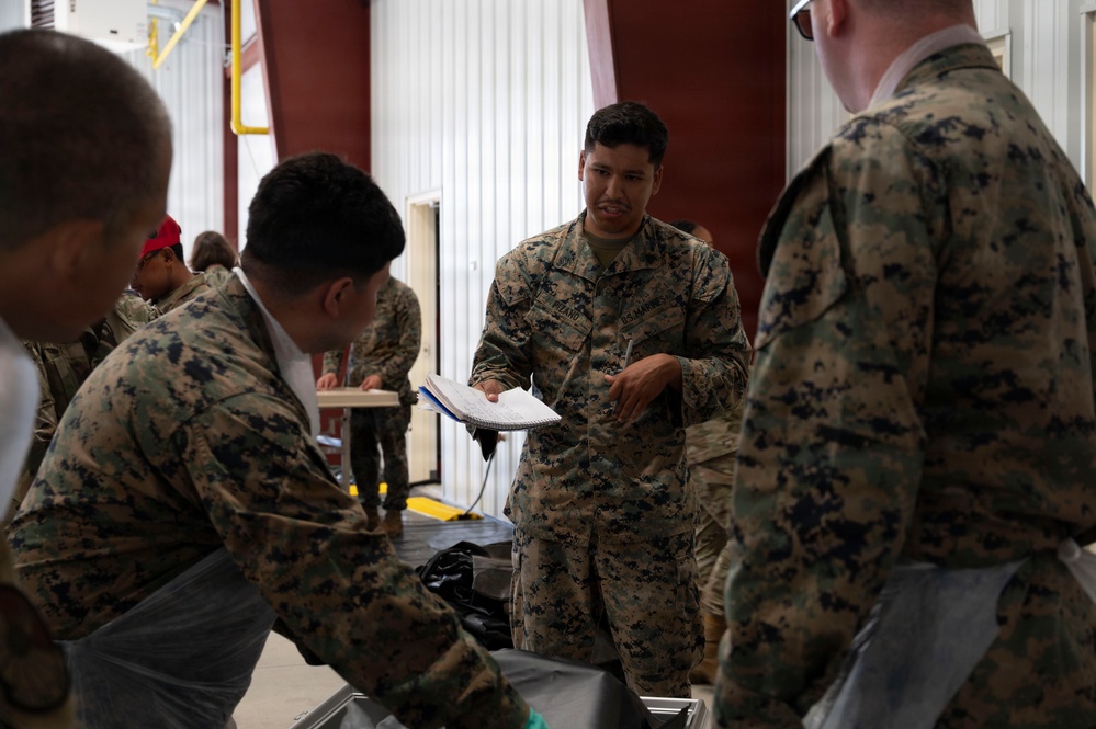 A group of U.S. Marines in MARPAT camouflage uniforms are gathered inside what appears to be a hangar or warehouse facility. A central Marine is holding a notebook or clipboard and appears to be conducting an inventory check or briefing. Multiple personnel are engaged in handling equipment or gear in the background.