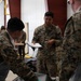 A group of U.S. Marines in MARPAT camouflage uniforms are gathered inside what appears to be a hangar or warehouse facility. A central Marine is holding a notebook or clipboard and appears to be conducting an inventory check or briefing. Multiple personnel are engaged in handling equipment or gear in the background.