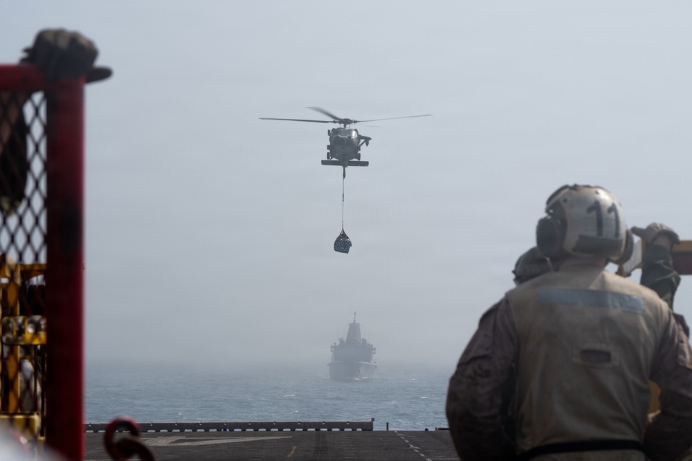 A military helicopter is conducting a vertical replenishment (VERTREP) operation, carrying a cargo net sling load over open ocean. Personnel on what appears to be a naval vessel's deck are observing the operation, wearing flight deck gear including helmets. A second naval vessel is visible in the hazy background.