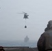 A military helicopter is conducting a vertical replenishment (VERTREP) operation, carrying a cargo net sling load over open ocean. Personnel on what appears to be a naval vessel's deck are observing the operation, wearing flight deck gear including helmets. A second naval vessel is visible in the hazy background.