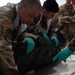 Military personnel in mixed camouflage uniforms (OCP/Army and MARPAT/Marine) are working together at a table, handling a black body bag while wearing green nitrile gloves and gray aprons. The scene appears to be a mortuary affairs or remains handling training exercise. Additional uniformed personnel observe in the background.
