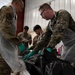Military personnel in mixed uniforms (Army OCP and Marine MARPAT camouflage) are conducting what appears to be a mortuary affairs or mass casualty training exercise. They are wearing protective aprons and green nitrile gloves while handling a black body bag on a table. Additional personnel in the background appear to be taking notes or documenting the procedure.