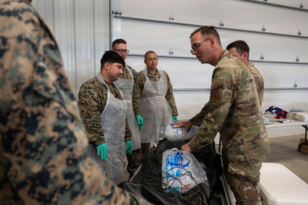 Military personnel in a hangar or warehouse setting are gathered around a table during what appears to be a food preparation or distribution activity. Several service members wearing protective aprons and green gloves are being instructed or briefed by a senior service member in OCP (Operational Camouflage Pattern) uniform. The presence of packaged food items and a cooler suggests a food service or humanitarian aid operation.