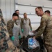 Military personnel in a hangar or warehouse setting are gathered around a table during what appears to be a food preparation or distribution activity. Several service members wearing protective aprons and green gloves are being instructed or briefed by a senior service member in OCP (Operational Camouflage Pattern) uniform. The presence of packaged food items and a cooler suggests a food service or humanitarian aid operation.