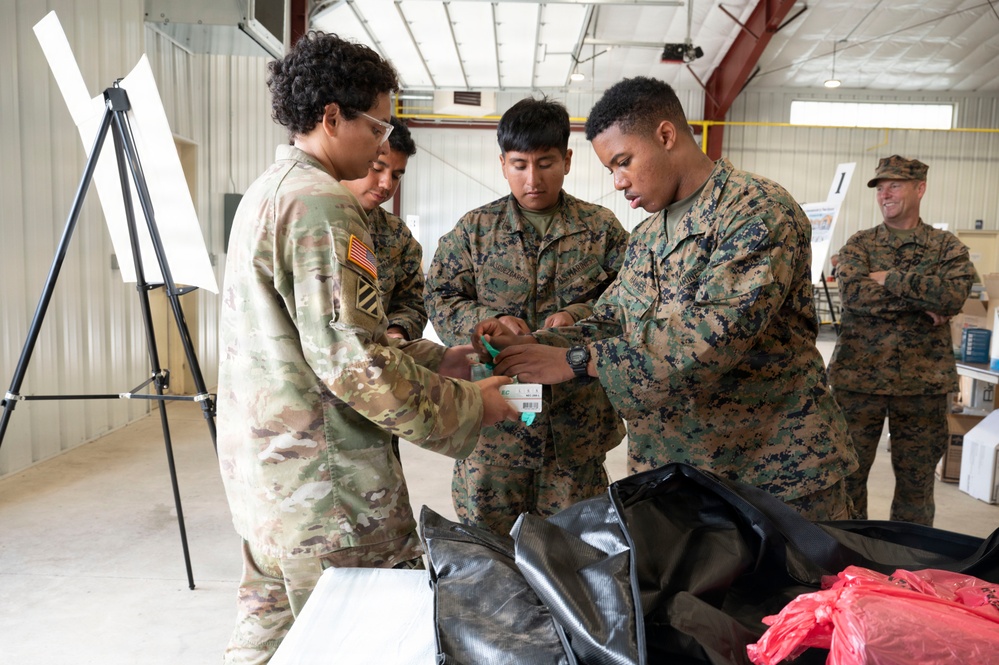 Military personnel from multiple branches are gathered inside what appears to be a training facility or warehouse. Three service members in the foreground are closely examining or handling a small item, while a fourth service member observes from the background with arms crossed. The scene suggests a training exercise or equipment inspection activity.