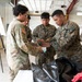 Military personnel from multiple branches are gathered inside what appears to be a training facility or warehouse. Three service members in the foreground are closely examining or handling a small item, while a fourth service member observes from the background with arms crossed. The scene suggests a training exercise or equipment inspection activity.