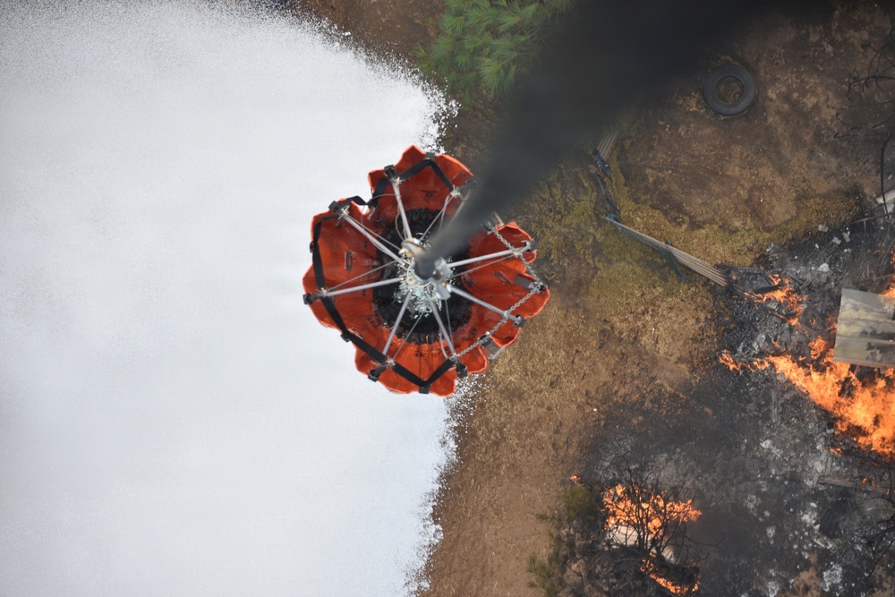 An aerial photograph taken from above shows a helicopter water bucket (Bambi bucket) actively releasing water or fire retardant onto a ground fire. The orange collapsible bucket is suspended by cables and is mid-discharge, with white foam/water spraying outward. Active flames, charred ground, debris, and a discarded tire are visible on the ground below.