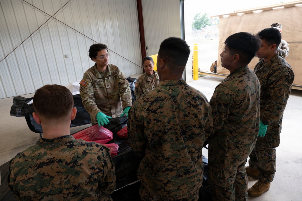 A U.S. Army soldier wearing OCP (Operational Camouflage Pattern) uniform is instructing a group of U.S. Marines in MARPAT uniforms inside what appears to be a warehouse or hangar facility. The Army instructor, wearing green protective gloves, appears to be conducting a briefing or demonstration involving black bags and red-packaged materials on a table. The setting suggests a training exercise or logistics/mortuary affairs operation.