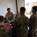 A U.S. Army soldier wearing OCP (Operational Camouflage Pattern) uniform is instructing a group of U.S. Marines in MARPAT uniforms inside what appears to be a warehouse or hangar facility. The Army instructor, wearing green protective gloves, appears to be conducting a briefing or demonstration involving black bags and red-packaged materials on a table. The setting suggests a training exercise or logistics/mortuary affairs operation.