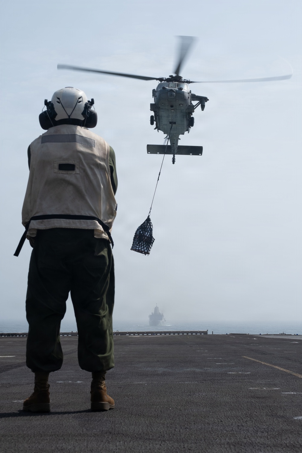A naval deck crew member stands on a ship's flight deck observing a military helicopter conducting a vertical replenishment (VERTREP) operation, with a cargo net suspended beneath the aircraft. The helicopter is approaching or hovering over the flight deck to deliver supplies. A second vessel is faintly visible in the background through hazy conditions.