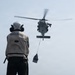 A naval deck crew member stands on a ship's flight deck observing a military helicopter conducting a vertical replenishment (VERTREP) operation, with a cargo net suspended beneath the aircraft. The helicopter is approaching or hovering over the flight deck to deliver supplies. A second vessel is faintly visible in the background through hazy conditions.