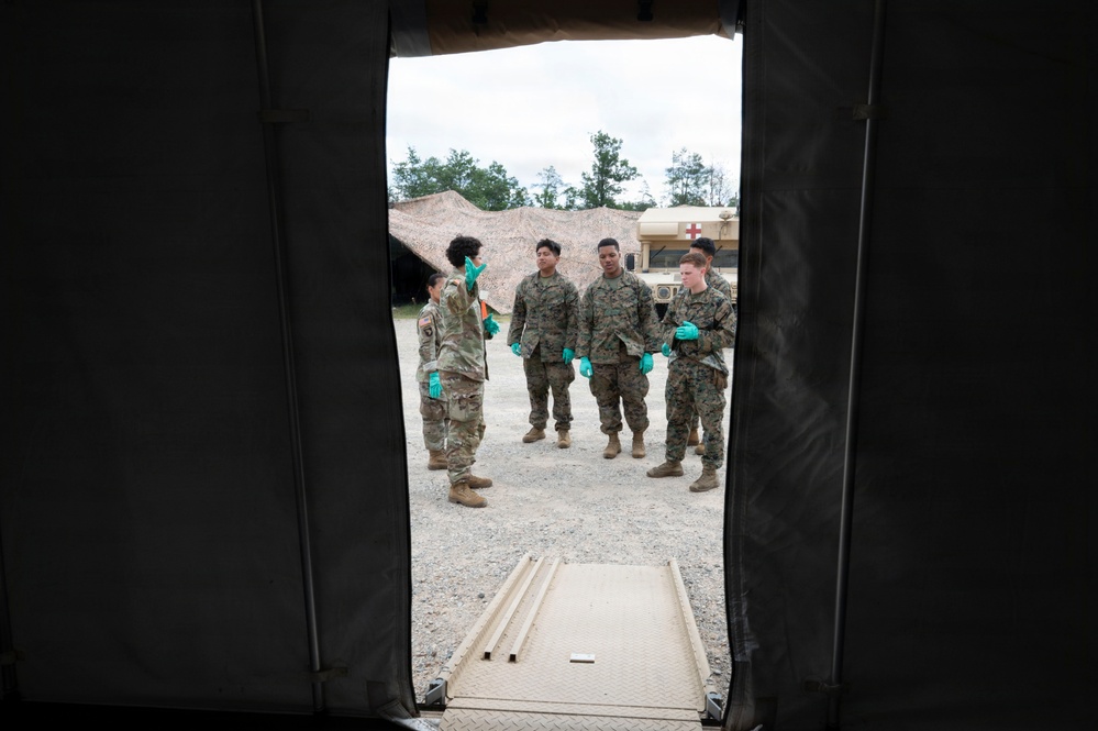 The image is taken from inside a military medical tent looking outward through an open doorway. Five military personnel in camouflage uniforms and teal/green medical gloves are gathered outside, with one individual appearing to brief or instruct the others. A military medical vehicle (HMMWV with red cross markings) and a camouflage-netted structure are visible in the background.