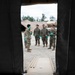 The image is taken from inside a military medical tent looking outward through an open doorway. Five military personnel in camouflage uniforms and teal/green medical gloves are gathered outside, with one individual appearing to brief or instruct the others. A military medical vehicle (HMMWV with red cross markings) and a camouflage-netted structure are visible in the background.