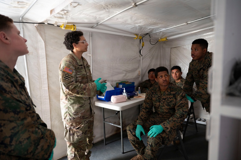 A U.S. Army soldier wearing protective eyewear and green latex gloves appears to be conducting a medical briefing or training session inside a military field tent. Several Marines in MARPAT camouflage uniforms, also wearing green latex gloves, are seated and listening attentively. The setting appears to be a field medical station with a supply table visible in the background.
