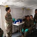 A U.S. Army soldier wearing protective eyewear and green latex gloves appears to be conducting a medical briefing or training session inside a military field tent. Several Marines in MARPAT camouflage uniforms, also wearing green latex gloves, are seated and listening attentively. The setting appears to be a field medical station with a supply table visible in the background.