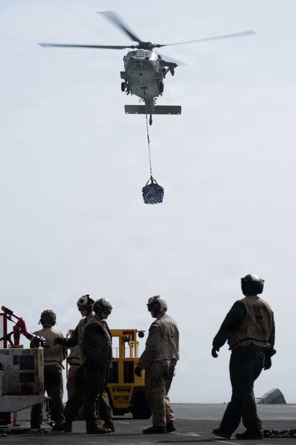 A military helicopter, appearing to be an MH-60 Seahawk variant, hovers above a ship's deck performing a vertical replenishment (VERTREP) operation, lowering a cargo net with supplies. Several personnel wearing helmets and protective vests are positioned on the deck, with a yellow forklift visible in the background ready to receive the cargo. The scene depicts a standard naval logistics resupply operation at sea.