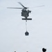 A military helicopter, appearing to be an MH-60 Seahawk variant, hovers above a ship's deck performing a vertical replenishment (VERTREP) operation, lowering a cargo net with supplies. Several personnel wearing helmets and protective vests are positioned on the deck, with a yellow forklift visible in the background ready to receive the cargo. The scene depicts a standard naval logistics resupply operation at sea.
