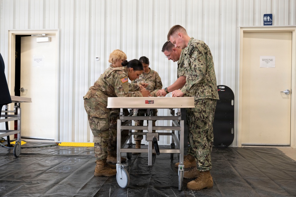 A group of military personnel from different branches are gathered around a medical gurney or examination table inside what appears to be a temporary or field facility. Three individuals wearing U.S. Army OCP (Operational Camouflage Pattern) uniforms and one wearing a U.S. Navy NWU (Naval Working Uniform) Type III are collaborating on a task at the table. The setting suggests a joint military medical training exercise or equipment setup.