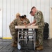 A group of military personnel from different branches are gathered around a medical gurney or examination table inside what appears to be a temporary or field facility. Three individuals wearing U.S. Army OCP (Operational Camouflage Pattern) uniforms and one wearing a U.S. Navy NWU (Naval Working Uniform) Type III are collaborating on a task at the table. The setting suggests a joint military medical training exercise or equipment setup.