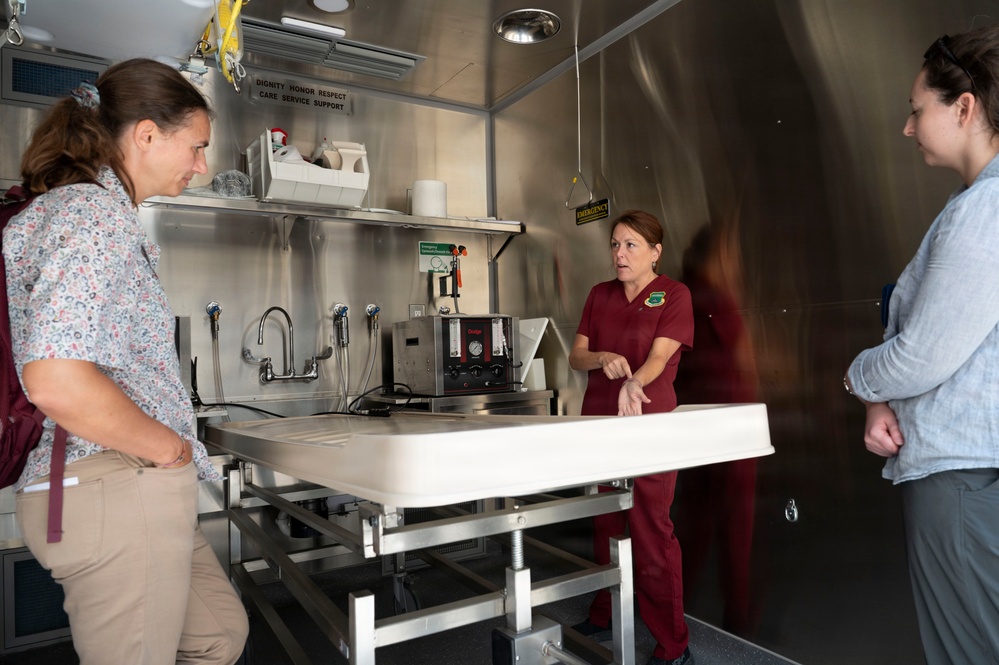 Three women are gathered inside what appears to be a mobile mortuary or medical examination unit, with one woman in maroon scrubs appearing to give a tour or briefing. The interior features stainless steel walls, a central examination/preparation table, sinks, and various medical/mortuary equipment. A sign on the wall reads 'DIGNITY HONOR RESPECT CARE SERVICE SUPPORT,' suggesting a military or official mortuary affairs context.