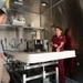 Three women are gathered inside what appears to be a mobile mortuary or medical examination unit, with one woman in maroon scrubs appearing to give a tour or briefing. The interior features stainless steel walls, a central examination/preparation table, sinks, and various medical/mortuary equipment. A sign on the wall reads 'DIGNITY HONOR RESPECT CARE SERVICE SUPPORT,' suggesting a military or official mortuary affairs context.