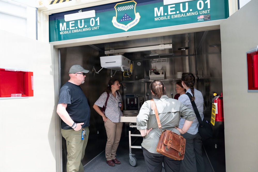 A group of four to five individuals are gathered at the entrance of an Air Force M.E.U. 01 (Mobile Embalming Unit), appearing to receive a briefing or tour of the facility. The unit displays the Air Force Mortuary Affairs Operations shield/crest on its signage. The interior shows stainless steel equipment, shelving, and a wheeled gurney or table.