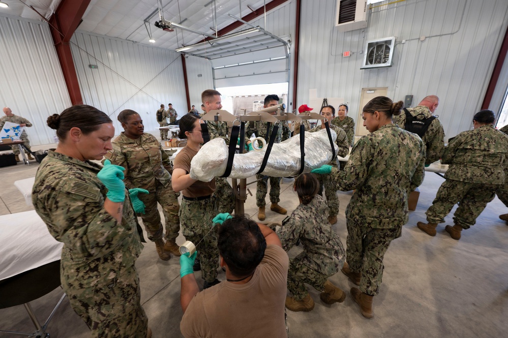 A group of military personnel in camouflage uniforms are gathered inside a large warehouse or hangar facility, working collaboratively around a stretcher or litter with a wrapped object on it. Several individuals are wearing green latex gloves, suggesting a medical or logistics preparation exercise. The scene appears to be a training exercise or disaster response preparation involving multiple service branches.