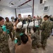 A group of military personnel in camouflage uniforms are gathered inside a large warehouse or hangar facility, working collaboratively around a stretcher or litter with a wrapped object on it. Several individuals are wearing green latex gloves, suggesting a medical or logistics preparation exercise. The scene appears to be a training exercise or disaster response preparation involving multiple service branches.
