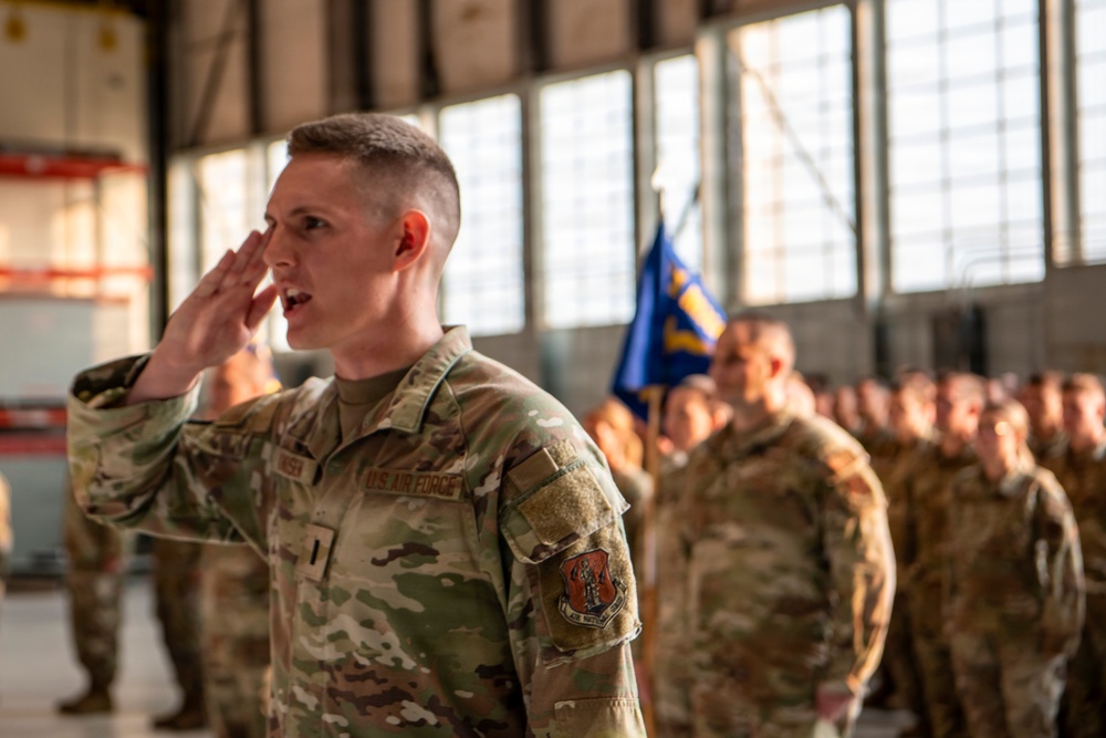 A U.S. Air Force service member in the foreground is rendering a salute during what appears to be a formal military ceremony held inside a hangar. The individual is wearing OCP (Operational Camouflage Pattern) uniform with 'U.S. AIR FORCE' tape and a unit patch visible on the sleeve. Behind them, rows of additional uniformed personnel are standing in formation with a blue military guidon flag visible in the background.