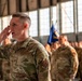 A U.S. Air Force service member in the foreground is rendering a salute during what appears to be a formal military ceremony held inside a hangar. The individual is wearing OCP (Operational Camouflage Pattern) uniform with 'U.S. AIR FORCE' tape and a unit patch visible on the sleeve. Behind them, rows of additional uniformed personnel are standing in formation with a blue military guidon flag visible in the background.