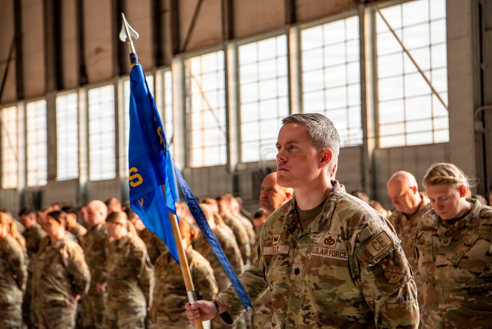 A U.S. Air Force military ceremony taking place inside a large hangar facility. A prominent airman in the foreground holds a blue unit guidon flag while wearing OCP (Operational Camouflage Pattern) uniform with 'U.S. AIR FORCE' and 'CE' (Civil Engineer) patches visible. Rows of uniformed airmen stand in formation in the background, with some appearing to have their heads bowed.