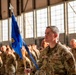 A U.S. Air Force military ceremony taking place inside a large hangar facility. A prominent airman in the foreground holds a blue unit guidon flag while wearing OCP (Operational Camouflage Pattern) uniform with 'U.S. AIR FORCE' and 'CE' (Civil Engineer) patches visible. Rows of uniformed airmen stand in formation in the background, with some appearing to have their heads bowed.
