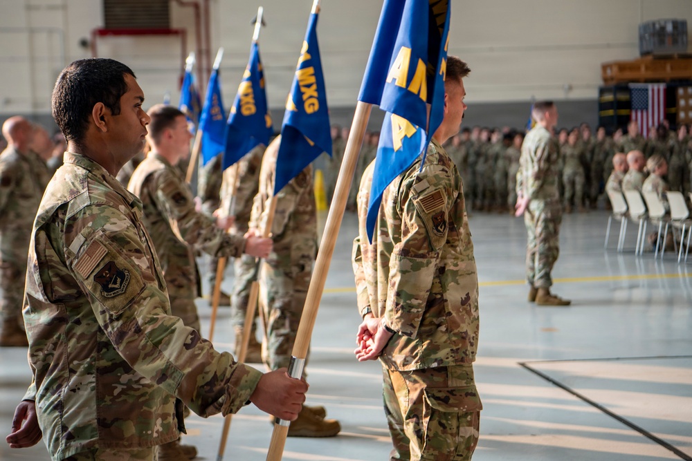 A formal military ceremony taking place inside an aircraft hangar, with uniformed personnel standing at attention in organized formations. Multiple guidon bearers are holding blue flags with yellow lettering, including visible 'MXG' (Maintenance Group) designations, suggesting a change of command or unit activation ceremony. An American flag is visible in the background along with rows of seated and standing personnel.