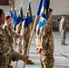 A formal military ceremony taking place inside an aircraft hangar, with uniformed personnel standing at attention in organized formations. Multiple guidon bearers are holding blue flags with yellow lettering, including visible 'MXG' (Maintenance Group) designations, suggesting a change of command or unit activation ceremony. An American flag is visible in the background along with rows of seated and standing personnel.