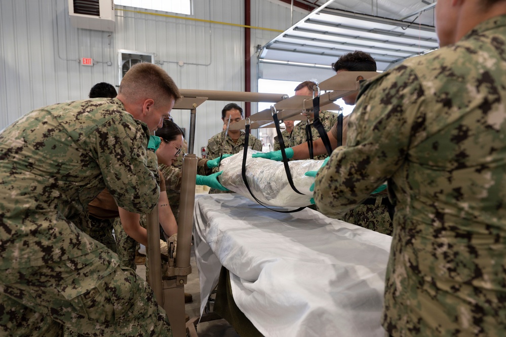 A group of military personnel in Navy Working Uniform (NWU) camouflage are conducting what appears to be a medical training exercise inside a warehouse or hangar facility. Multiple service members wearing green latex gloves are working together to transfer or position a wrapped/bundled patient or mannequin onto a medical gurney. The scene suggests a mass casualty or patient transfer training drill.