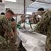 A group of military personnel in Navy Working Uniform (NWU) camouflage are conducting what appears to be a medical training exercise inside a warehouse or hangar facility. Multiple service members wearing green latex gloves are working together to transfer or position a wrapped/bundled patient or mannequin onto a medical gurney. The scene suggests a mass casualty or patient transfer training drill.