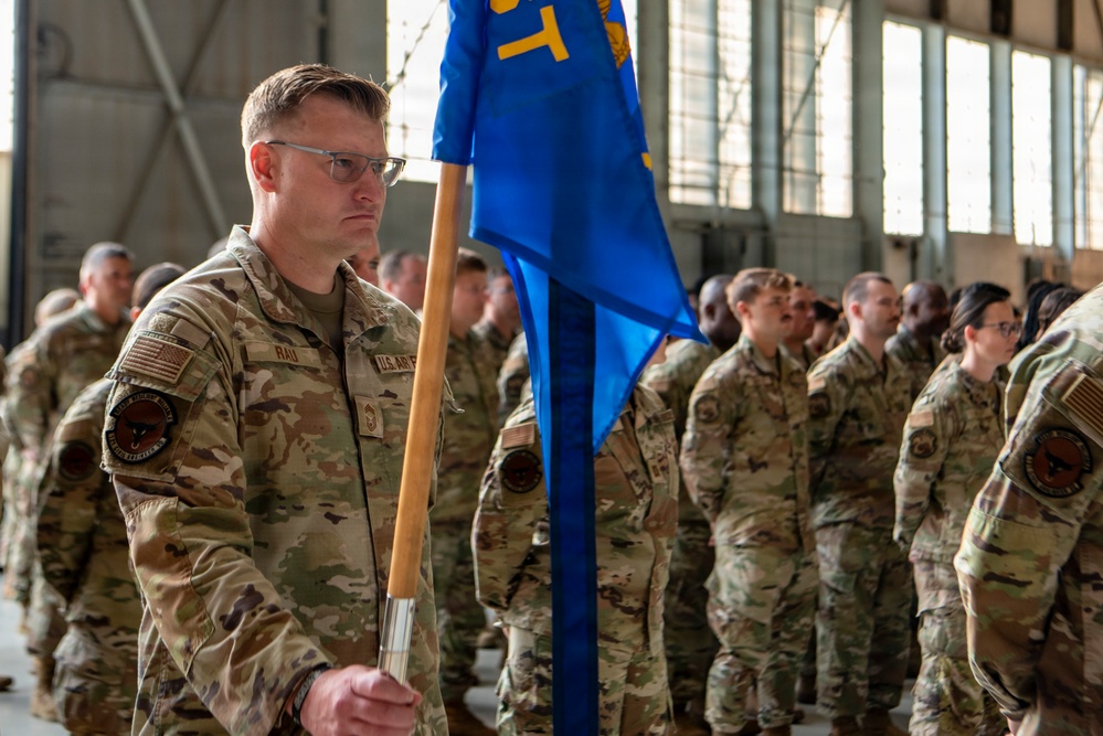 A U.S. Air Force military formation ceremony taking place inside a hangar. A senior non-commissioned officer identified by name tape as 'RAU' stands at the forefront holding a blue unit guidon flag with gold insignia. Multiple Airmen in OCP (Operational Camouflage Pattern) uniforms are standing at attention in formation behind him.